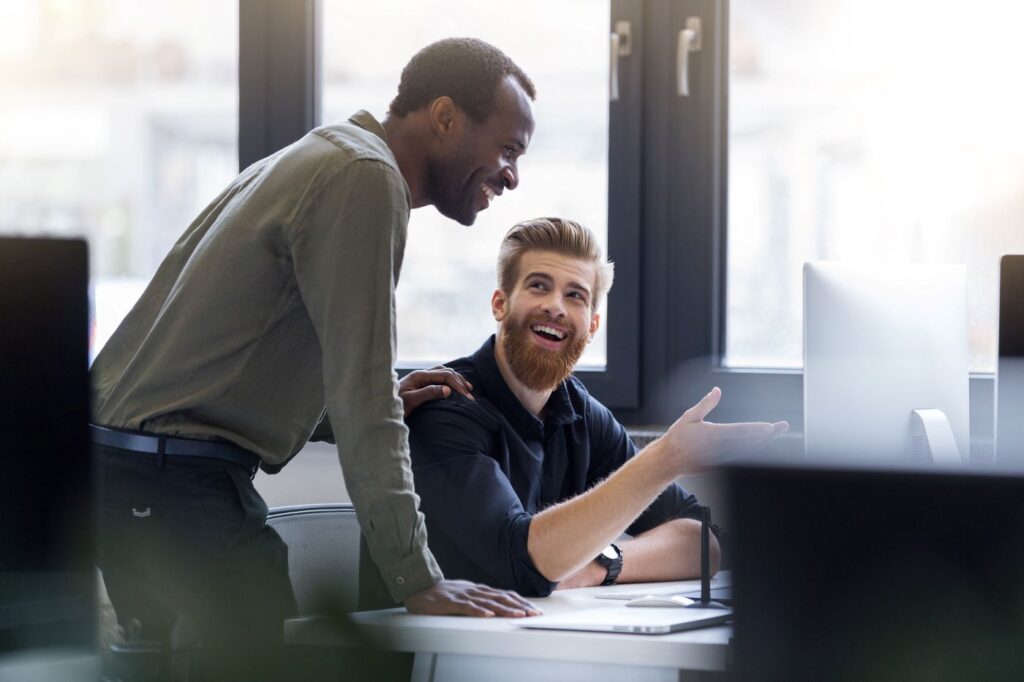 Office Management Zwei Männer unterhalten sich und betrachten lachend eine Computerbildschirm.